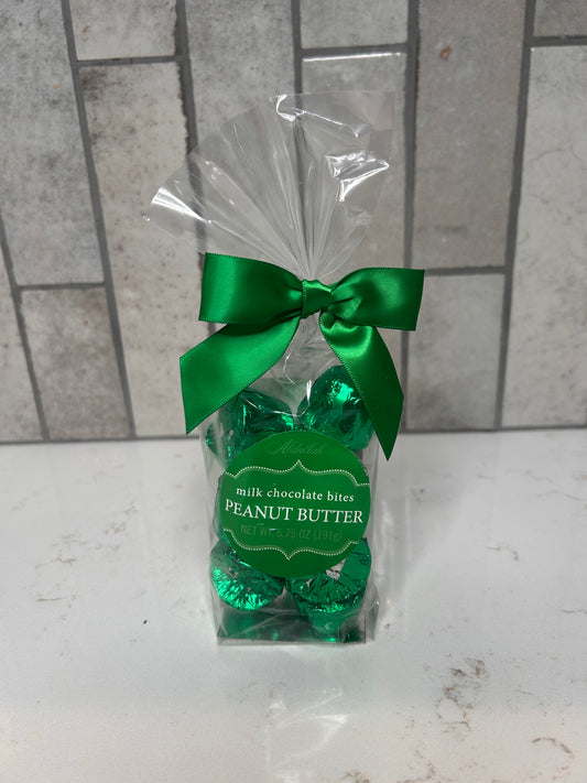 A clear gift bag filled with green-foil wrapped milk chocolate peanut butter bites, tied with a green satin bow, displayed on a white countertop with a gray tile background.