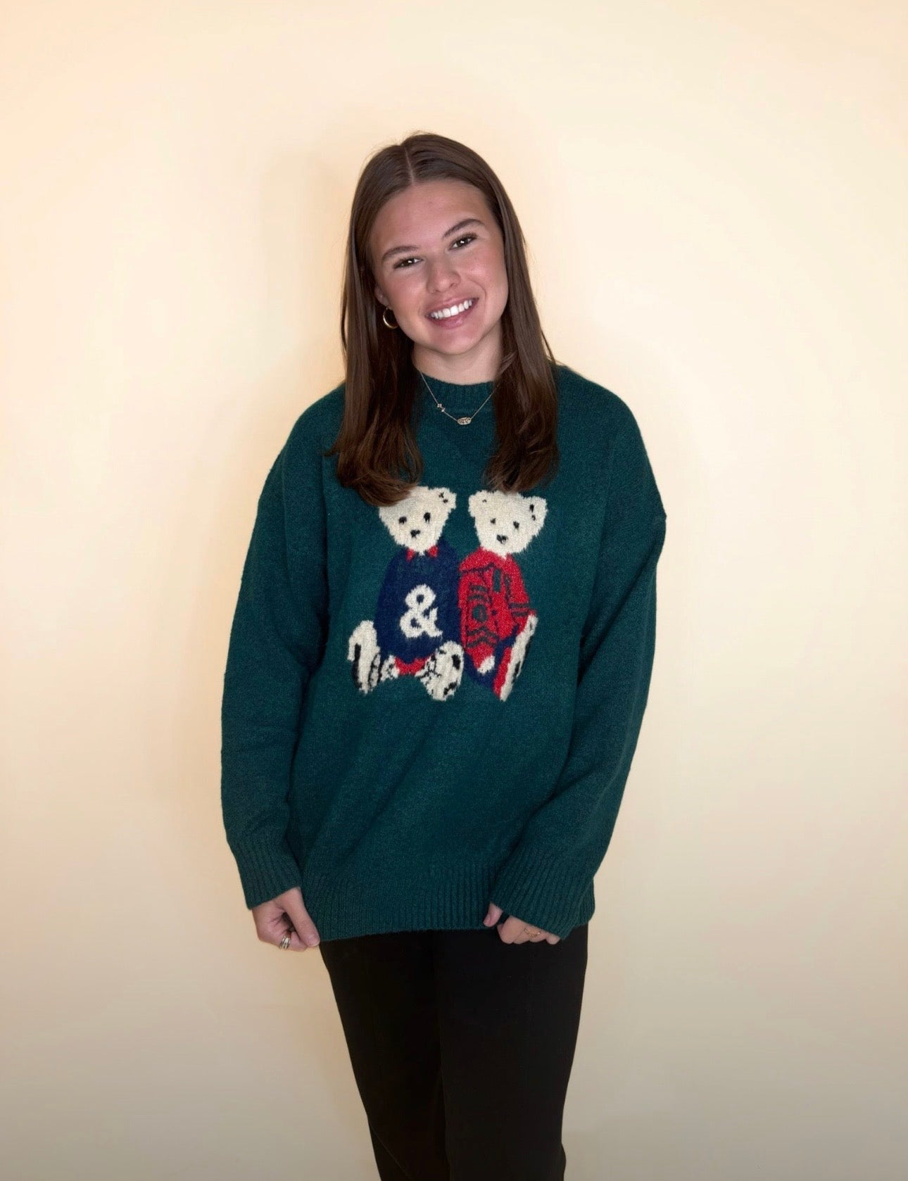 Woman wearing a forest-green oversized sweater with teddy bear graphics and black pants, smiling in front of a neutral background.