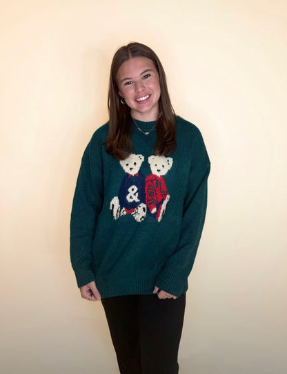 Woman wearing a forest-green oversized sweater with teddy bear graphics and black pants, smiling in front of a neutral background.