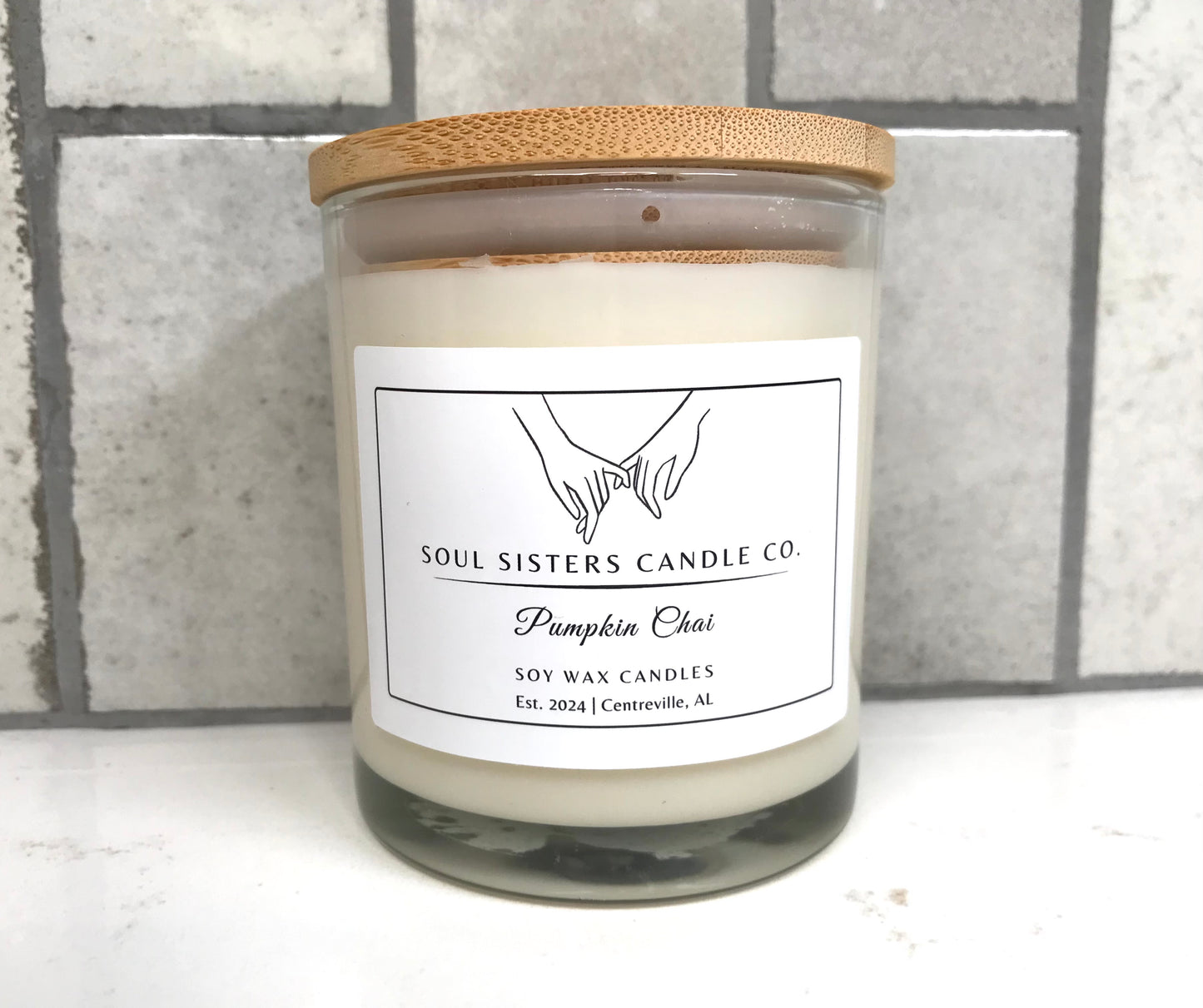 Scented candle in a glass jar with a wooden lid on a white surface with a gray tiled background