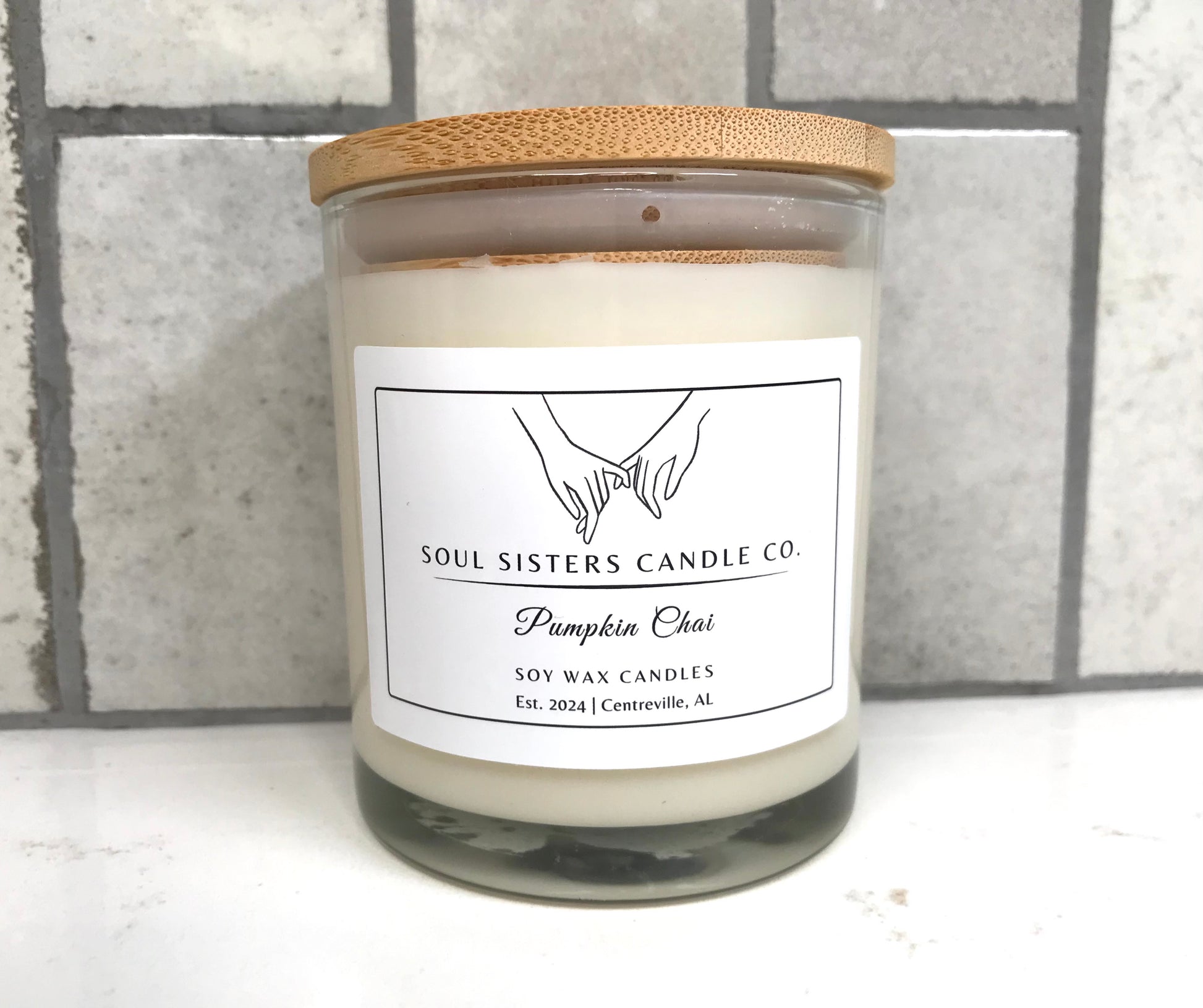 Scented candle in a glass jar with a wooden lid on a white surface with a gray tiled background