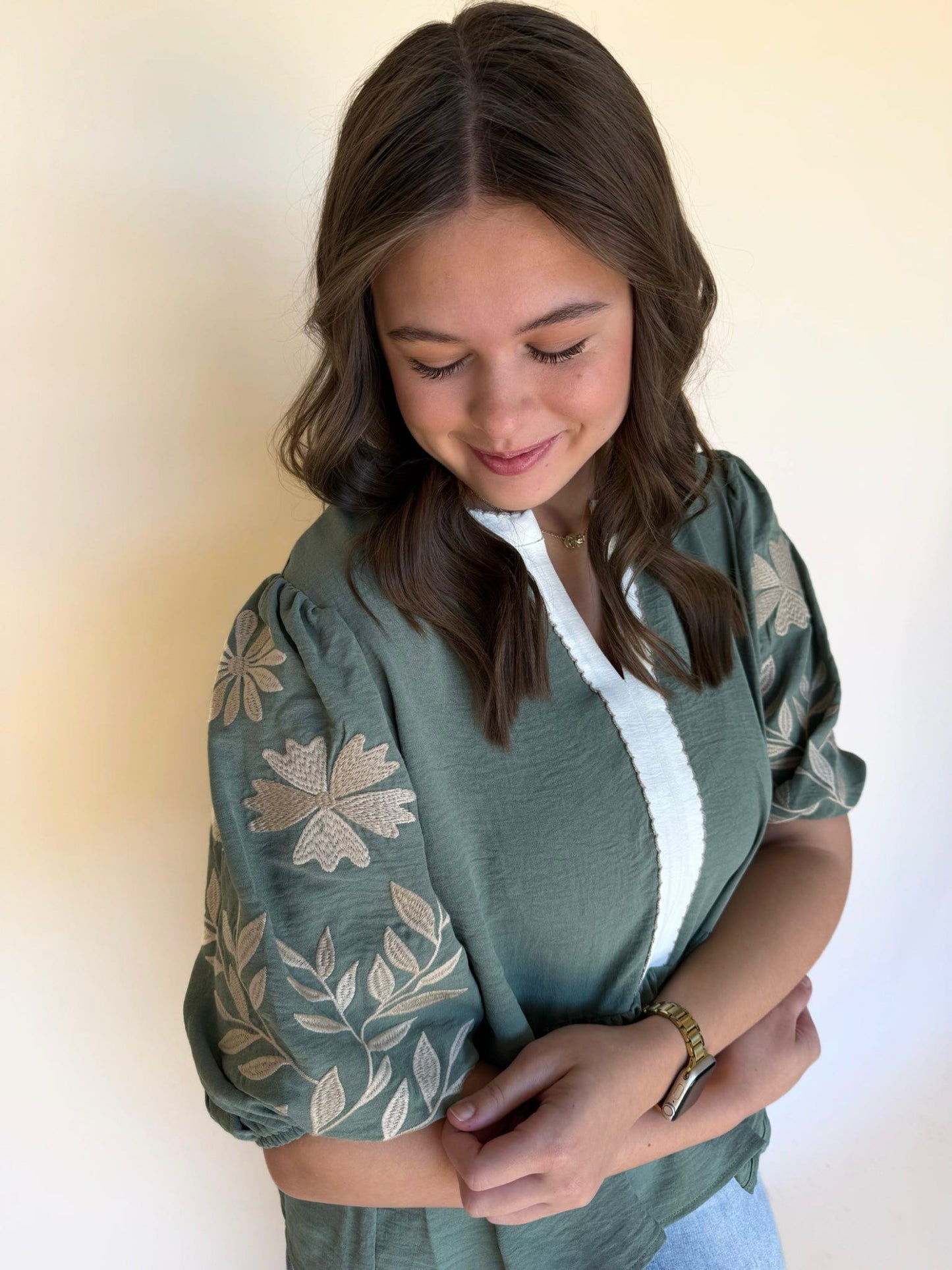 Close-up of a woman wearing a sage green embroidered blouse with puff sleeves and cream floral stitching, smiling softly while looking down.