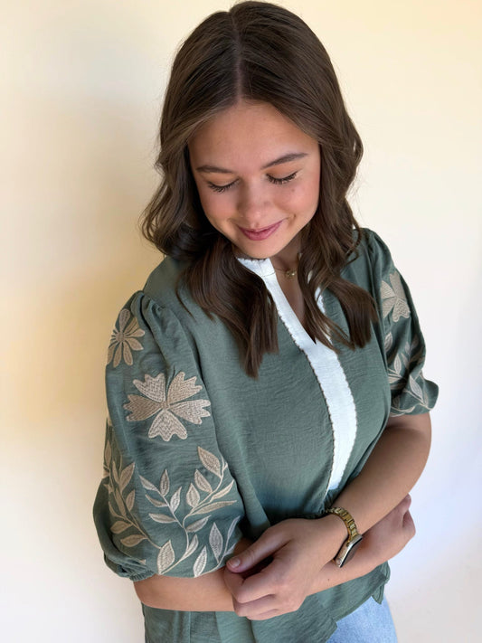 Close-up of a woman wearing a sage green embroidered blouse with puff sleeves and cream floral stitching, smiling softly while looking down.