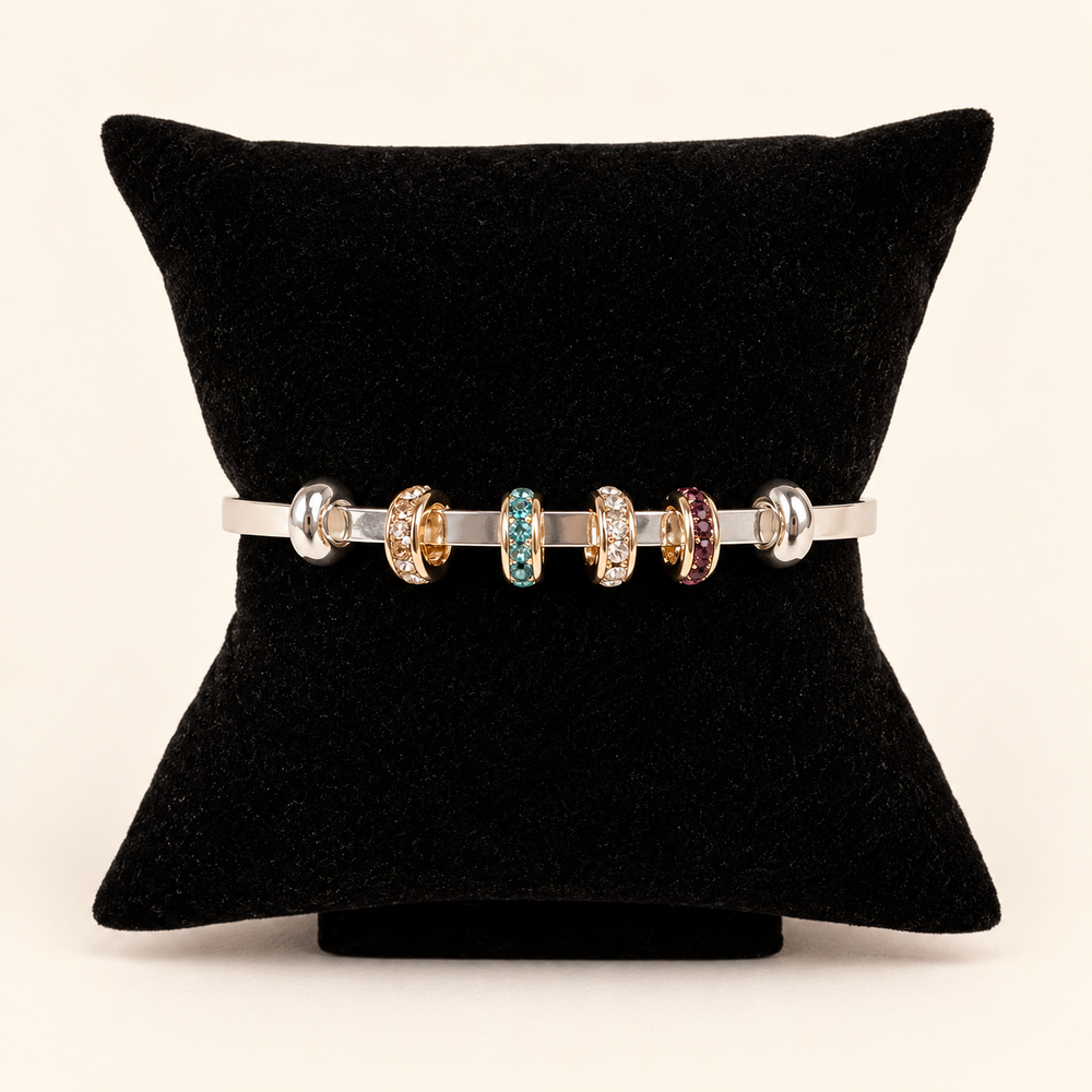 Silver open cuff bracelet styled with gold-tone crystal charm rings in clear, turquoise blue, and burgundy displayed on a black velvet pillow against a cream studio background.