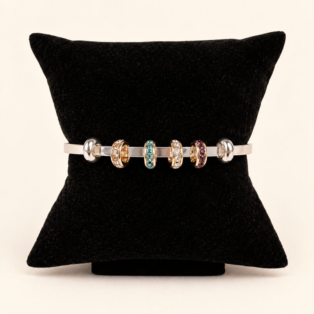 Silver open cuff bracelet styled with gold-tone crystal charm rings in clear, turquoise blue, and burgundy displayed on a black velvet pillow against a cream studio background.