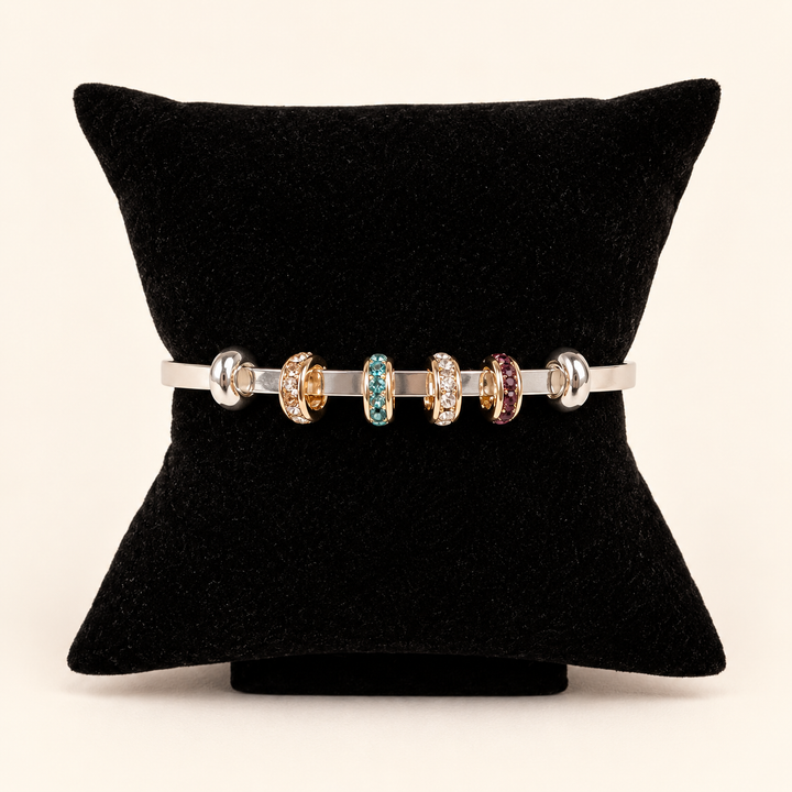 Silver open cuff bracelet styled with gold-tone crystal charm rings in clear, turquoise blue, and burgundy displayed on a black velvet pillow against a cream studio background.