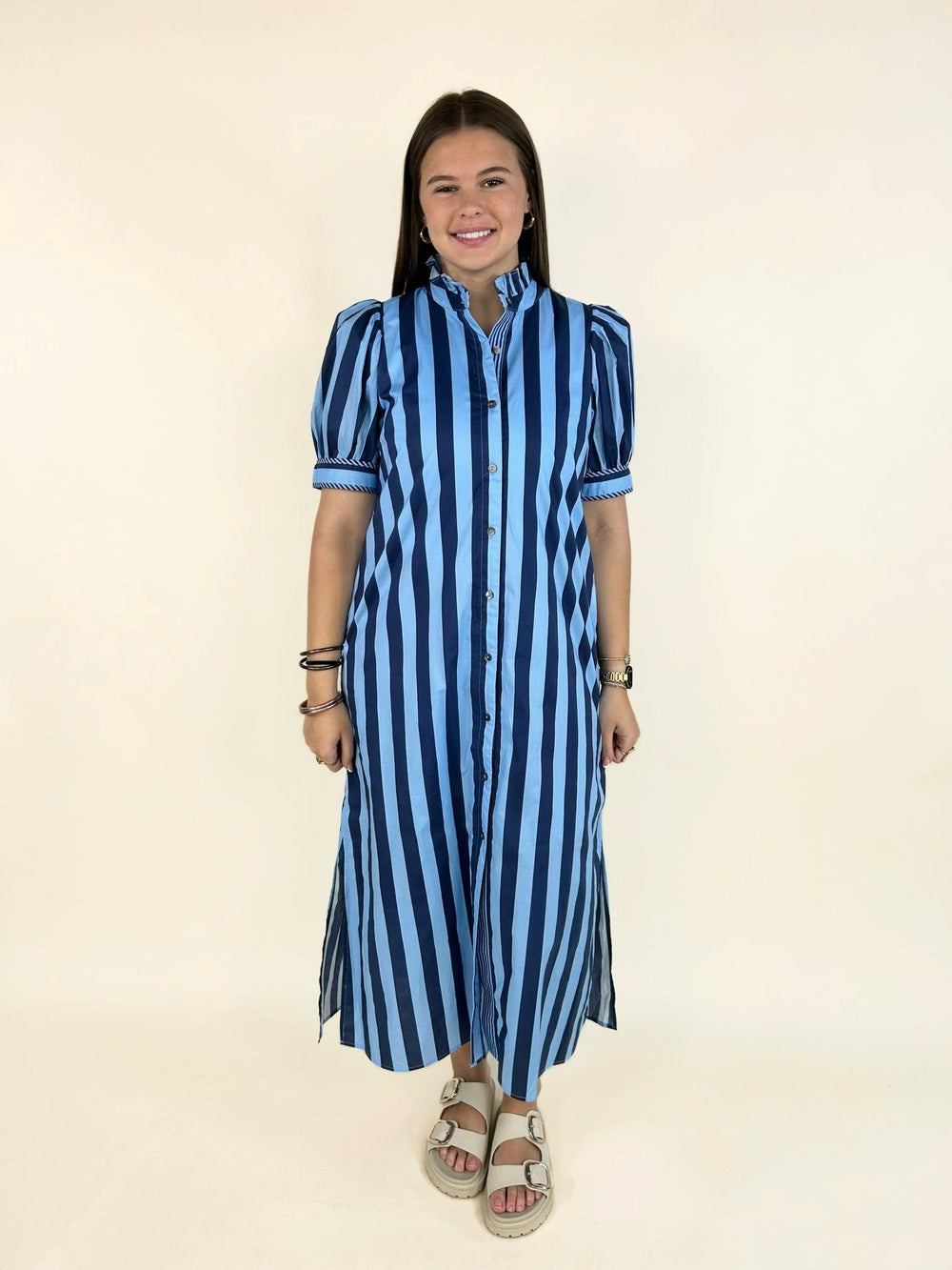 Model wearing a blue striped button-front midi shirt dress with a ruffle neckline, puff sleeves, side pockets, and side slits, styled with cream double-buckle sandals.