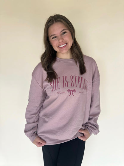 Person wearing a pink sweatshirt with 'She is Strong' text on a plain background