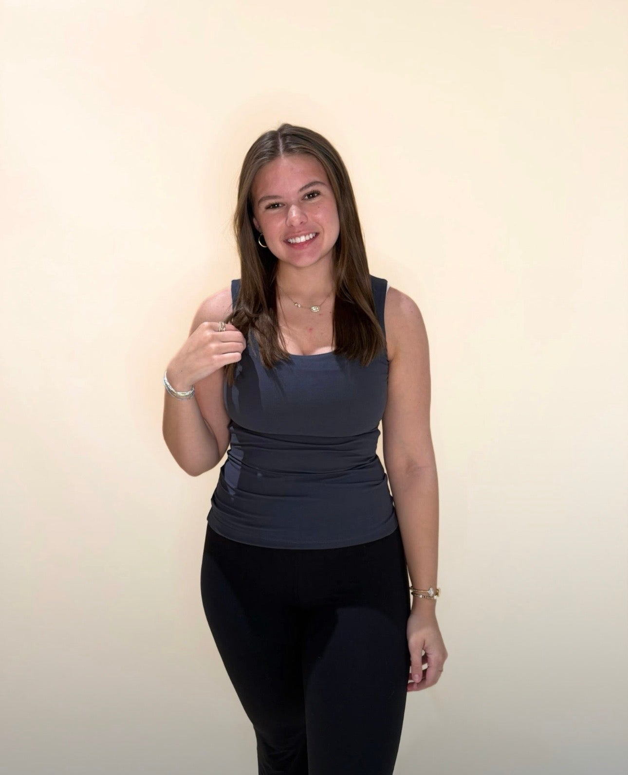 Young woman wearing a charcoal gray tank top paired with black leggings, standing and smiling in front of a cream backdrop.
