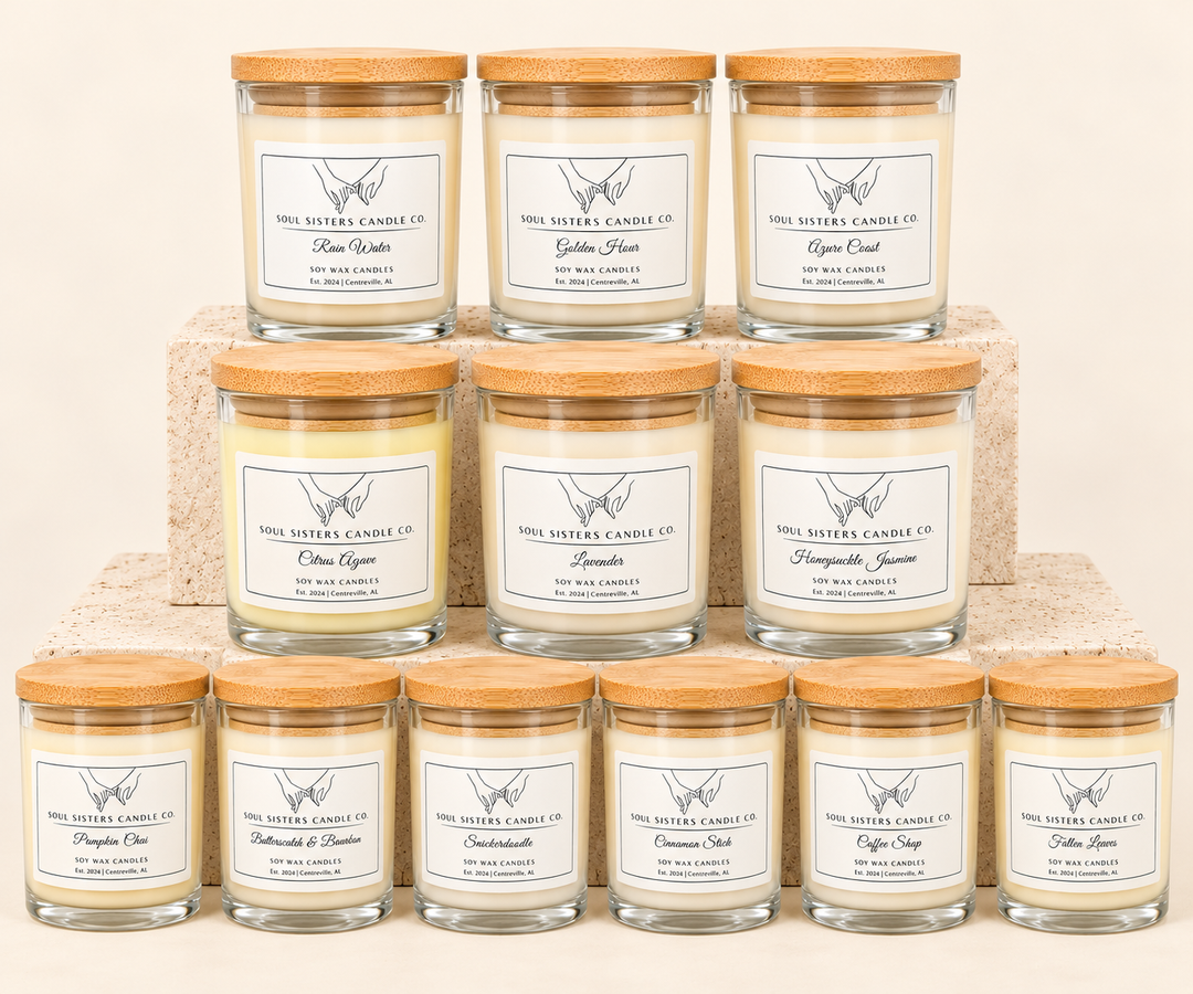 Soul Sisters Candle Co. soy wax candle collection featuring 12 scents, including Rain Water, Golden Hour, Azure Coast, Citrus Agave, Lavender, Honeysuckle Jasmine, Pumpkin Chai, Butterscotch &amp; Bourbon, Snickerdoodle, Cinnamon Stick, Coffee Shop, and Fallen Leaves, displayed in clear glass jars with natural bamboo lids on tiered stone risers against a cream studio background.