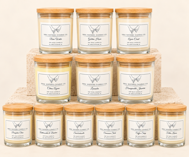Soul Sisters Candle Co. soy wax candle collection featuring 12 scents, including Rain Water, Golden Hour, Azure Coast, Citrus Agave, Lavender, Honeysuckle Jasmine, Pumpkin Chai, Butterscotch &amp; Bourbon, Snickerdoodle, Cinnamon Stick, Coffee Shop, and Fallen Leaves, displayed in clear glass jars with natural bamboo lids on tiered stone risers against a cream studio background.