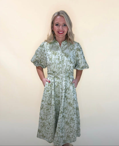 Woman wearing a light green floral print midi dress with puff sleeves and a tie waist, hands in pockets, smiling against a neutral cream background.