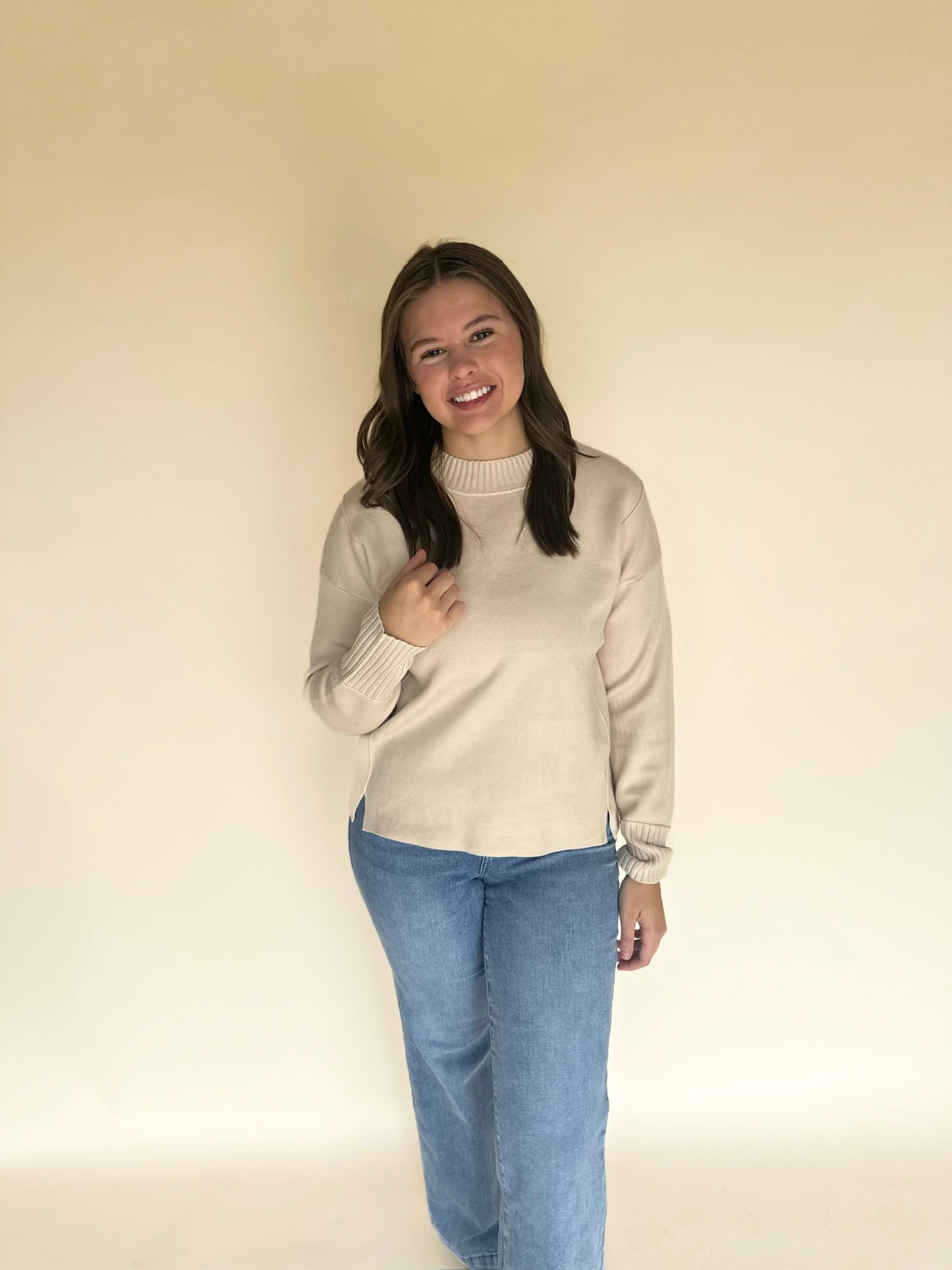 Woman wearing an oatmeal sweater and blue jeans against a plain background