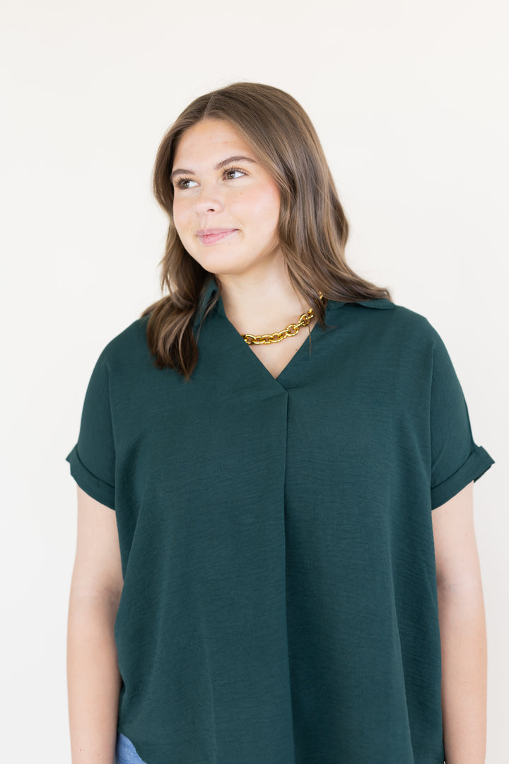 A woman wearing a dark green short-sleeve top with a V-neckline, photographed against a plain off-white background.
