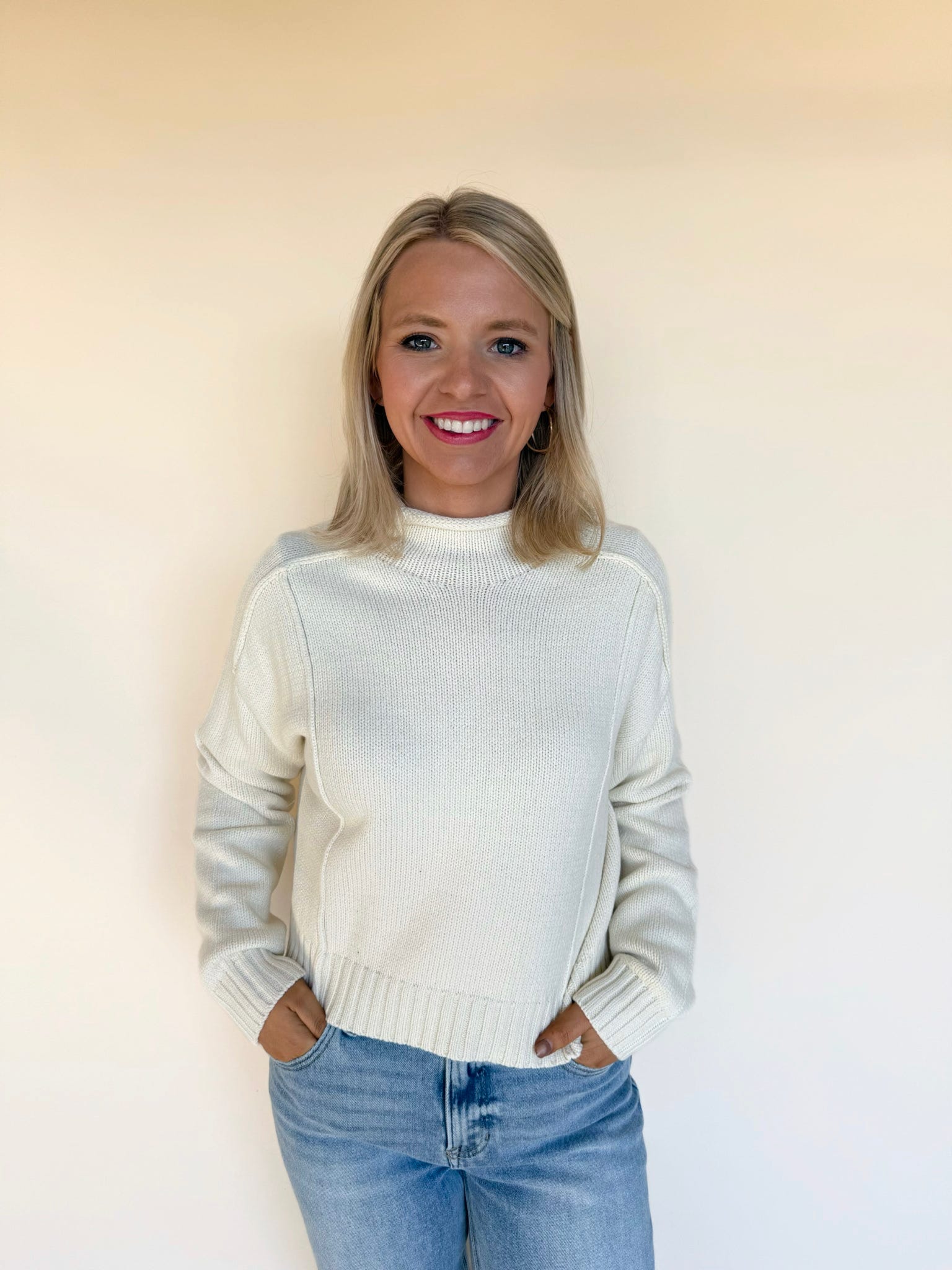 Woman wearing an ivory mock-neck knit sweater with ribbed cuffs and hem, styled with light-wash blue jeans against a neutral background.