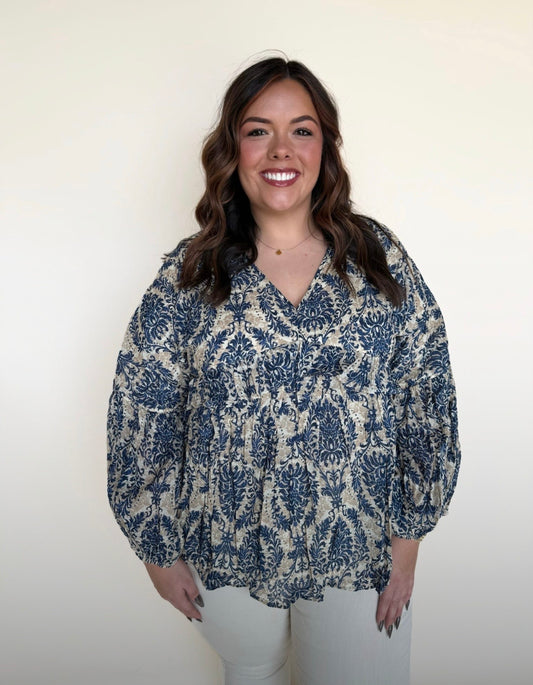 Plus-size model wearing a blue and beige damask print long-sleeve blouse with a flowy babydoll fit, V-neckline, and lightweight fabric — front view.