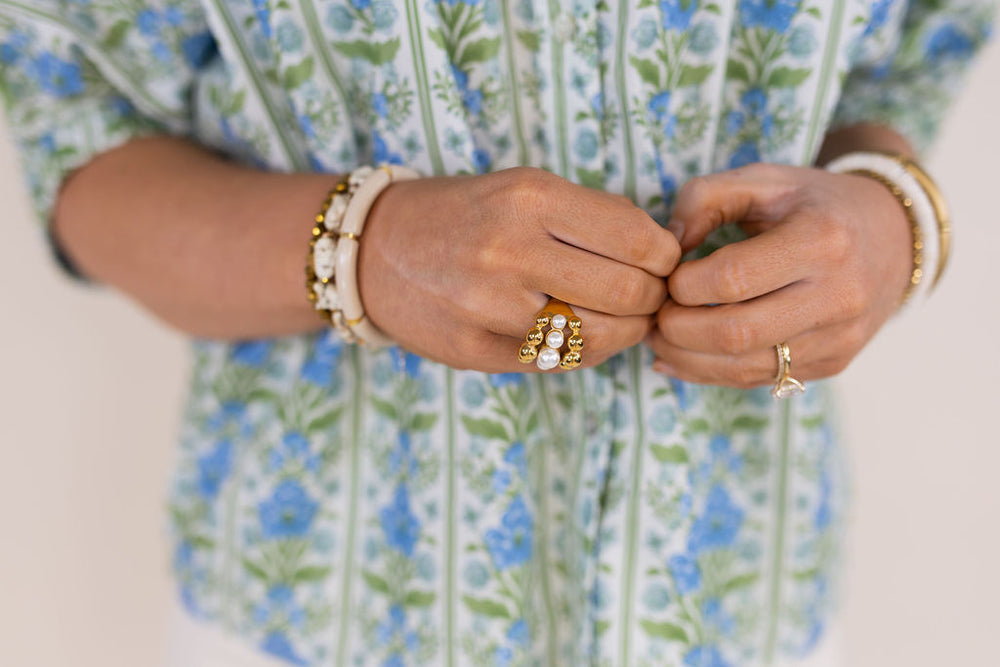 Close-up of a person's hands wearing multiple gold rings with white pearls, along with white beaded bracelets, while wearing a green and blue floral patterned top.