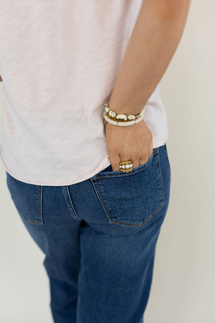A close-up lifestyle shot of a person wearing a white t-shirt, blue jeans, a gold ring, and a stack of white and gold bracelets, standing with their hand in their jeans pocket against a plain off-white background.