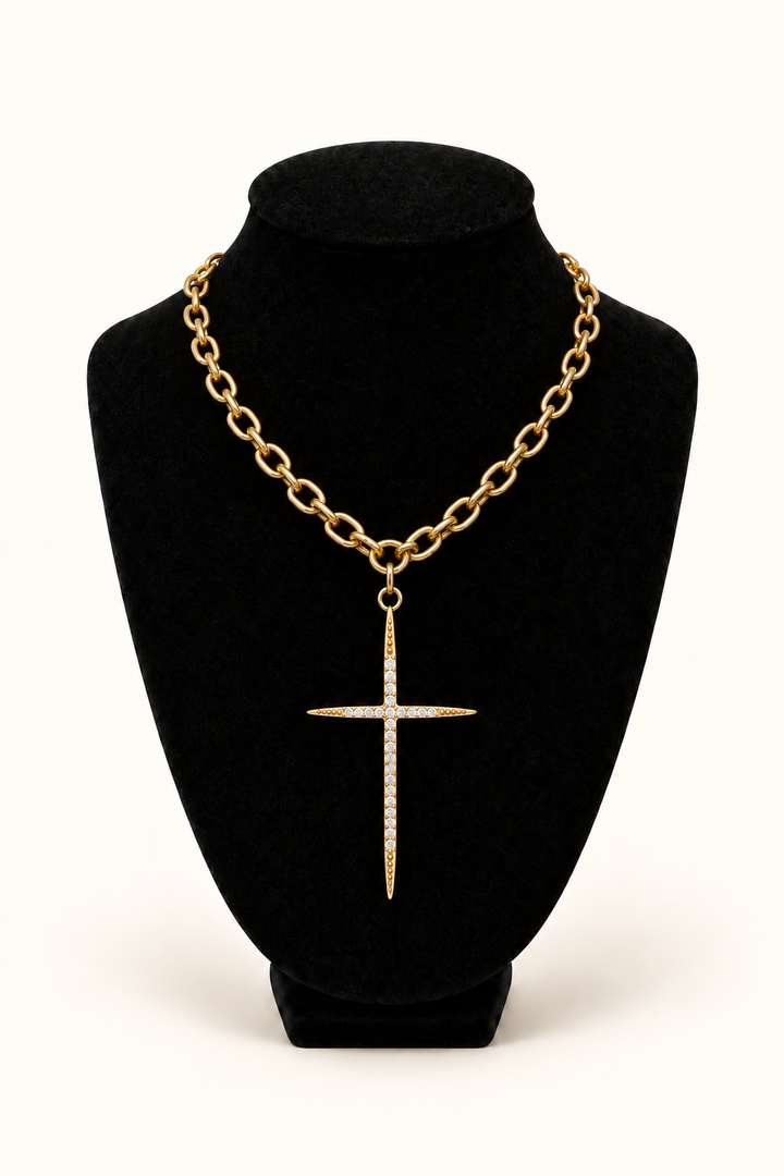 Gold open-link chain necklace with a long, slender cross pendant featuring clear crystal accents and dotted gold tips, displayed on a black velvet bust against a warm cream background.