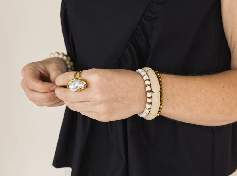 A close-up of a person's hands wearing multiple bracelets and a ring. The jewelry includes a gold ring with a large white pearl, a beaded bracelet, and stacked cream and gold bracelets, worn against a black top against a light beige background.
