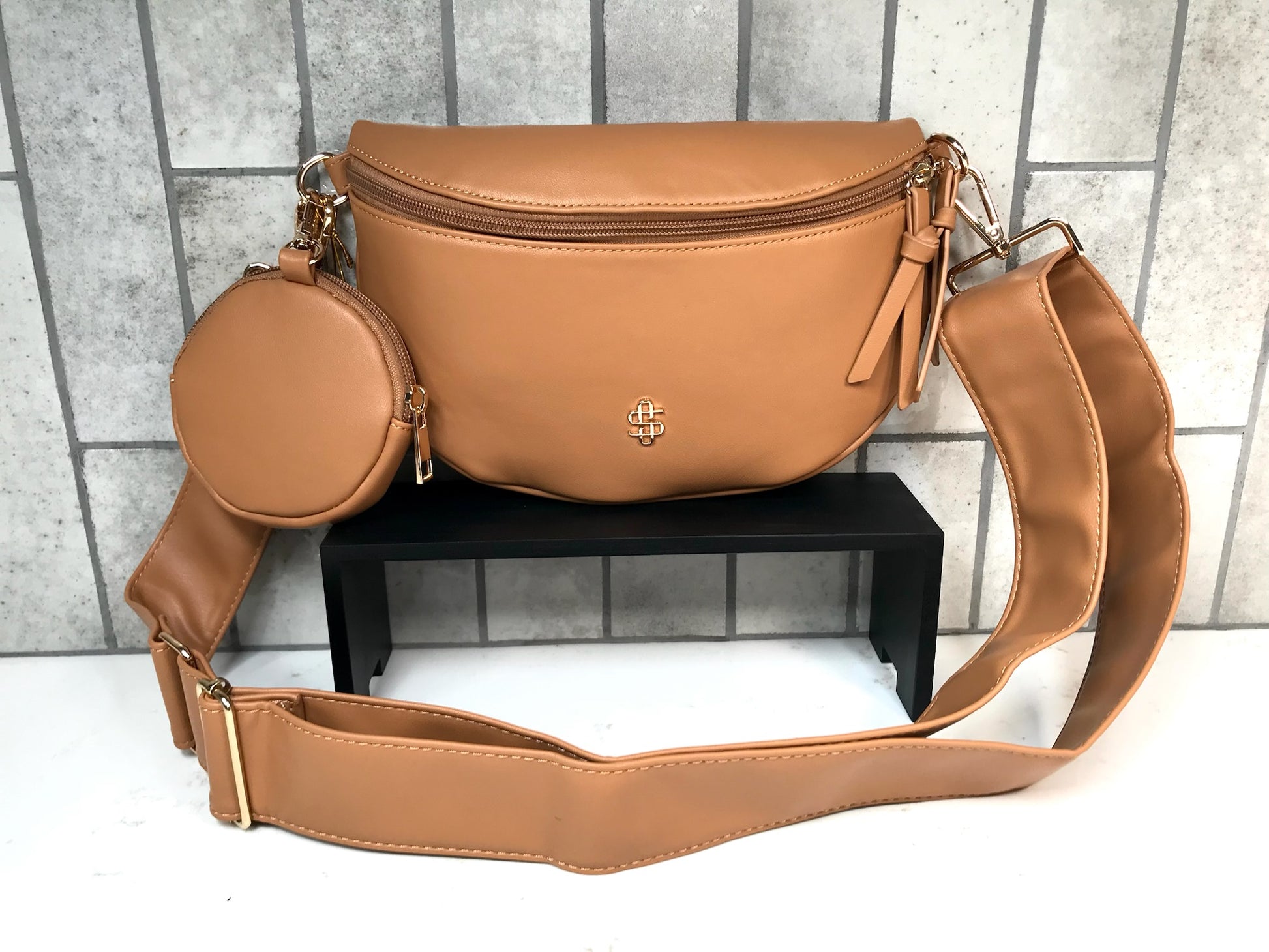 Tan leather crossbody bag with a smaller round bag on a black stand against a tiled wall background.