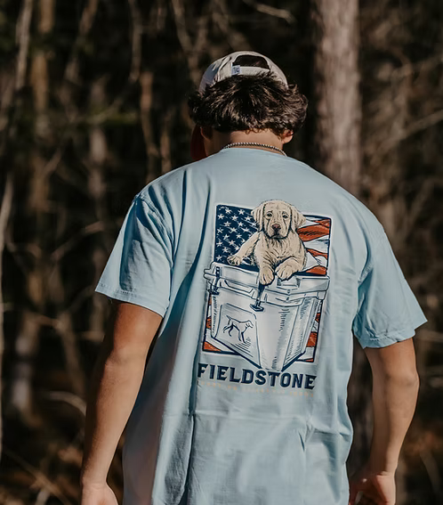 Fieldstone Sporting Lifestyle Brand light blue graphic t-shirt featuring Labrador retriever puppy sitting on cooler with American flag backdrop, Southern outdoor apparel, hunting dog tee, patriotic comfort shirt