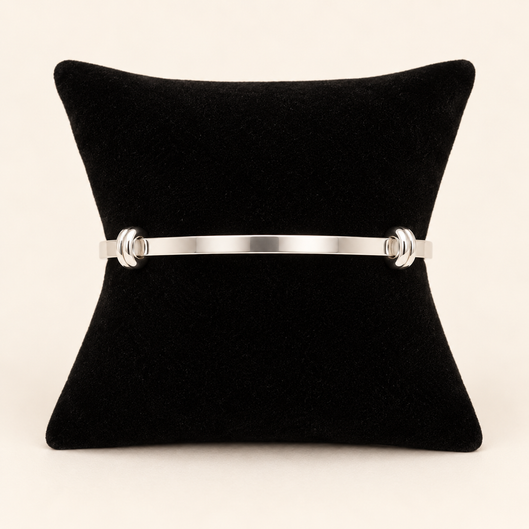 Silver open cuff bracelet with polished knot-style end details displayed on a black velvet pillow against a cream studio background.