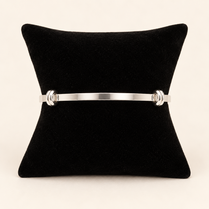 Silver open cuff bracelet with polished knot-style end details displayed on a black velvet pillow against a cream studio background.