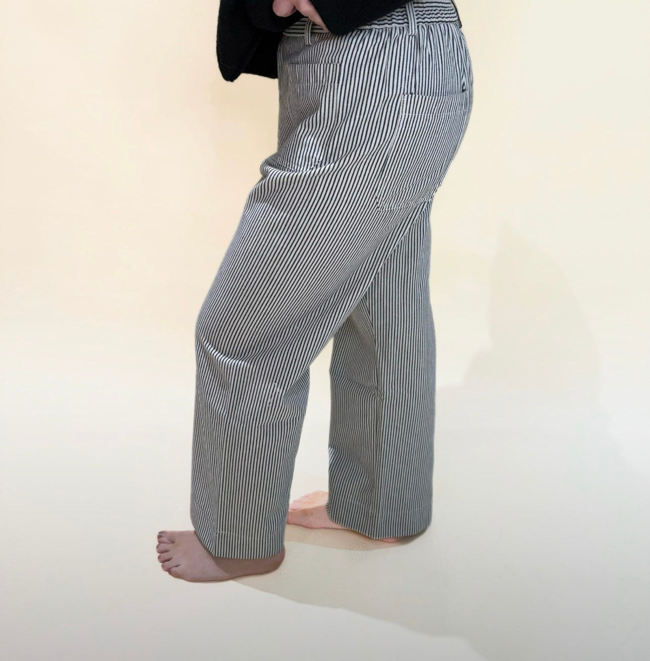 Side view of striped black and white straight-leg pants with relaxed fit and front pocket detail, shown on a barefoot model standing against a cream background.