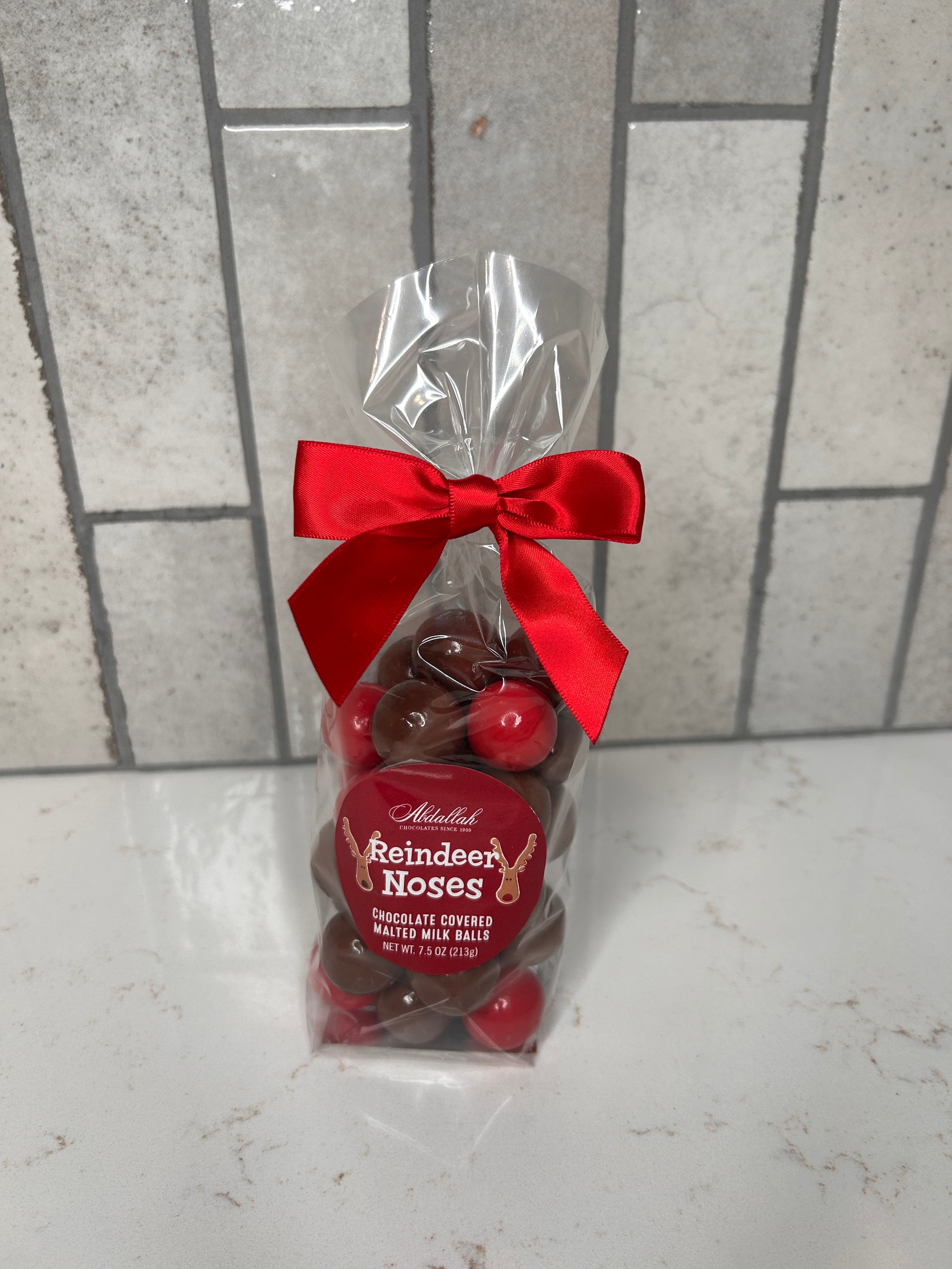 Clear gift bag of chocolate-covered malted milk balls labeled ‘Reindeer Noses,’ tied with a red satin bow, displayed on a light countertop with neutral tile backdrop.