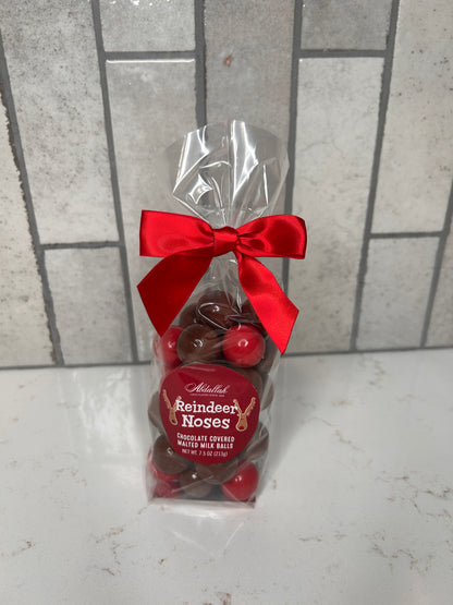 Clear gift bag of chocolate-covered malted milk balls labeled ‘Reindeer Noses,’ tied with a red satin bow, displayed on a light countertop with neutral tile backdrop.