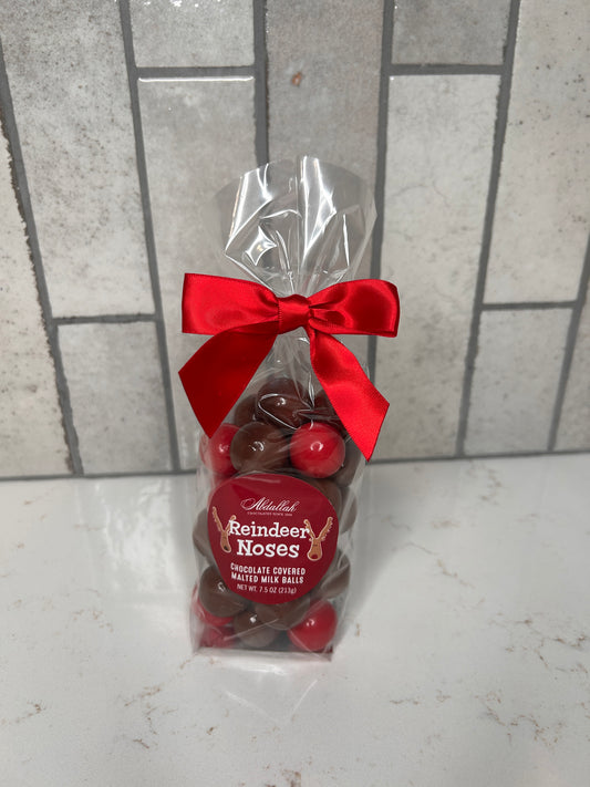Clear gift bag of chocolate-covered malted milk balls labeled ‘Reindeer Noses,’ tied with a red satin bow, displayed on a light countertop with neutral tile backdrop.