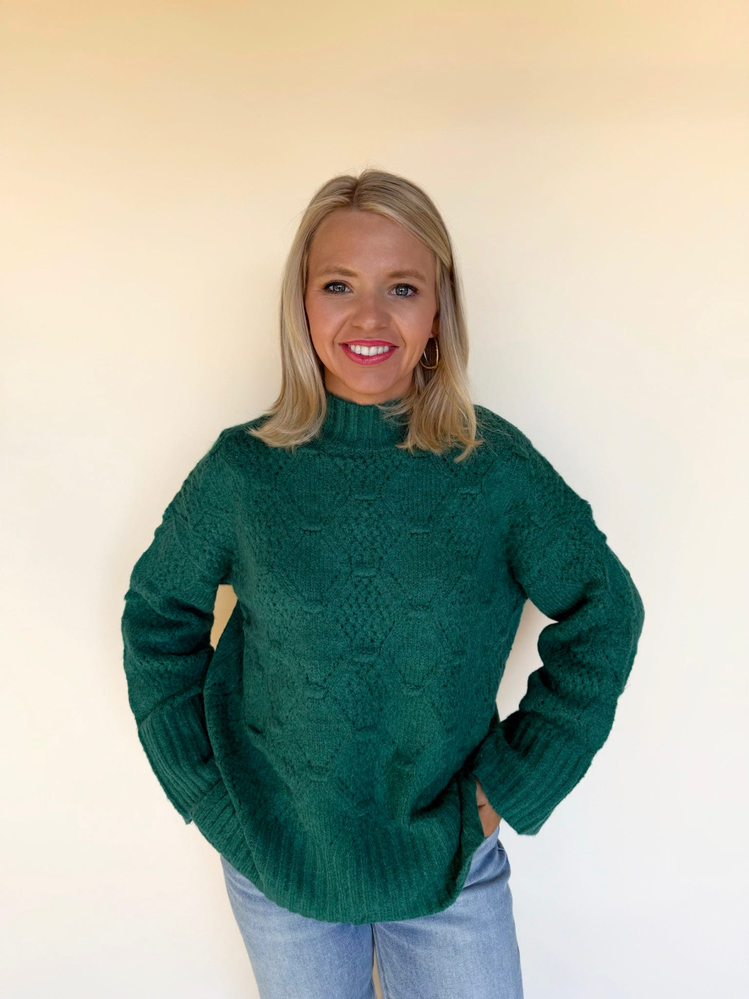 Woman wearing a green textured knit sweater with a mock neckline and long sleeves, paired with light-wash blue jeans against a neutral background.