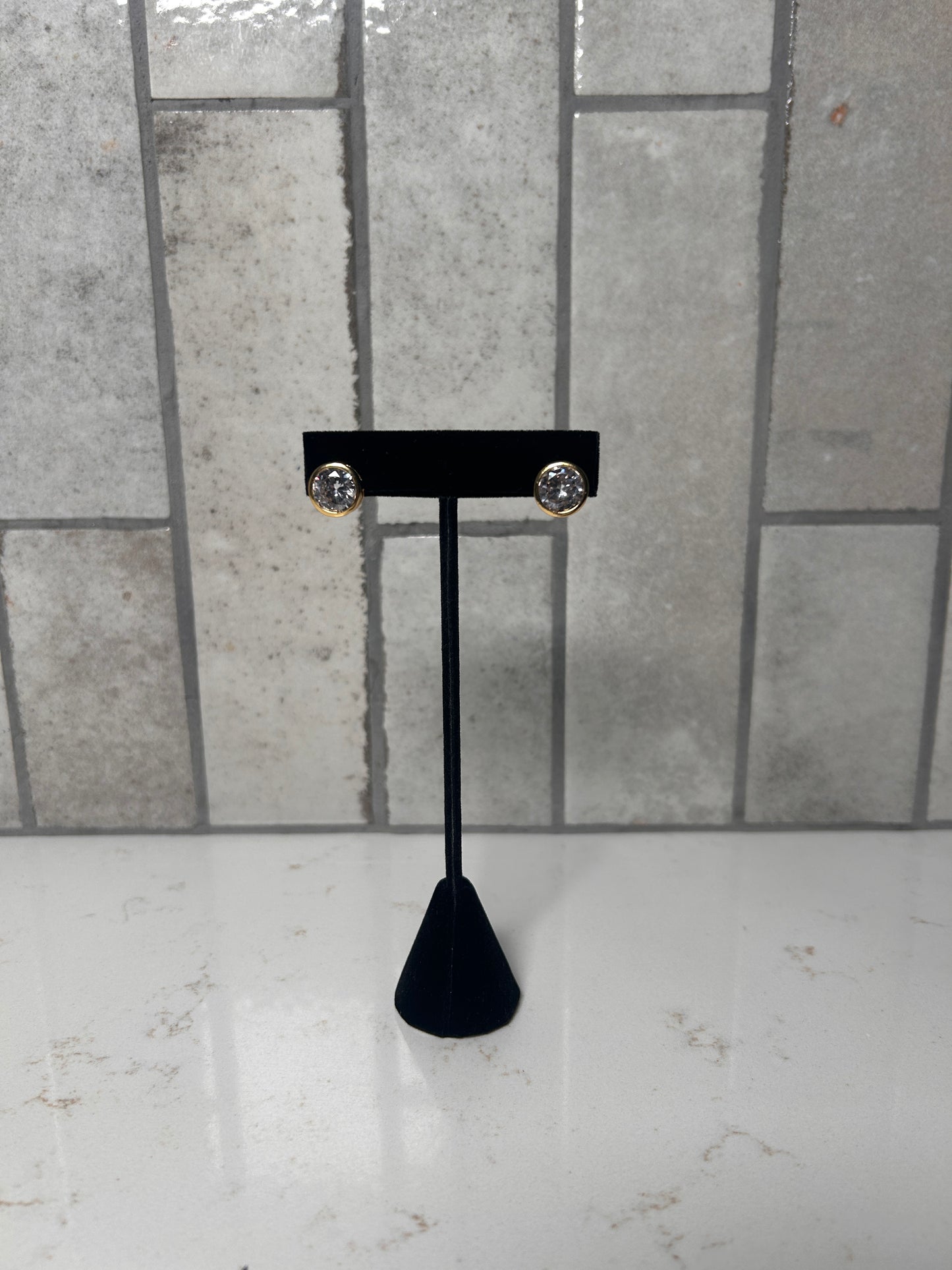 Gold bezel stud earrings with clear round crystal displayed on black jewelry stand against neutral tile background