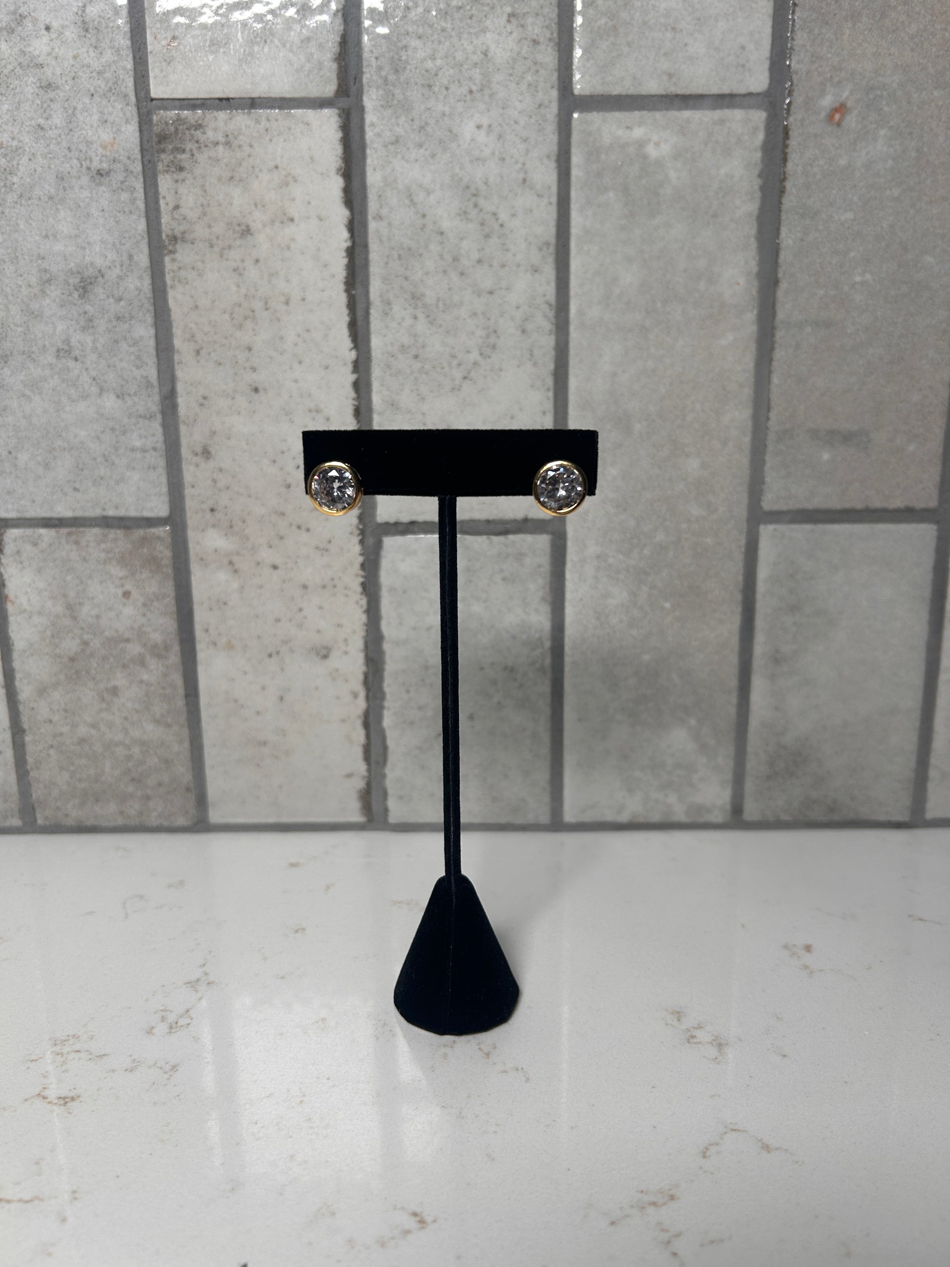 Gold bezel stud earrings with clear round crystal displayed on black jewelry stand against neutral tile background