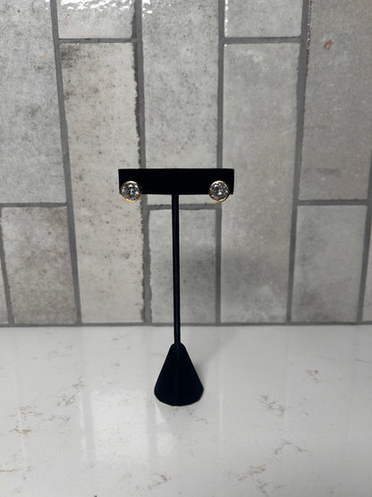 Gold bezel stud earrings with clear round crystal displayed on black jewelry stand against neutral tile background