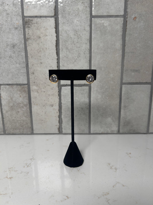 Gold bezel stud earrings with clear round crystal displayed on black jewelry stand against neutral tile background