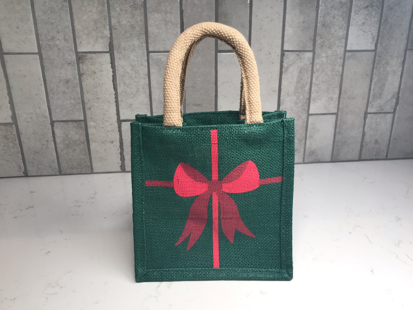 Christmas gift tote in dark green woven fabric with red bow print and beige handles, photographed against light gray tile backdrop.