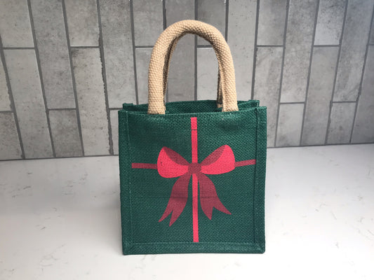 Christmas gift tote in dark green woven fabric with red bow print and beige handles, photographed against light gray tile backdrop.