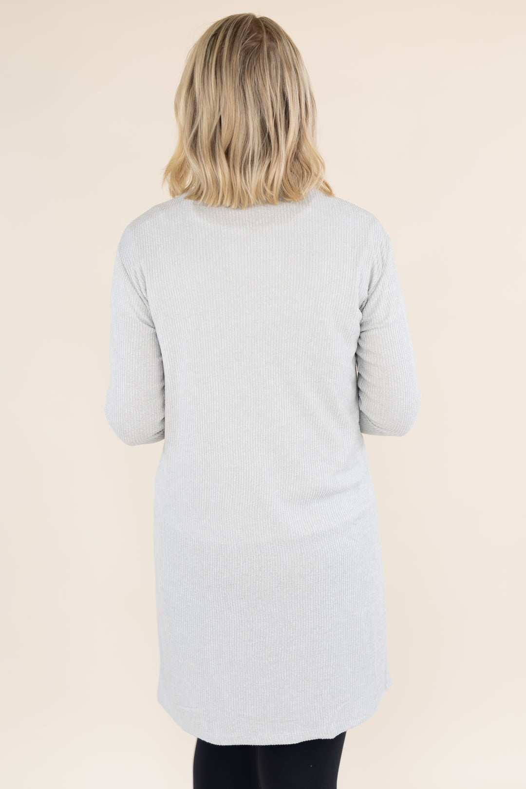 A woman viewed from behind wearing a light gray ribbed knit long-sleeve tunic top, set against a solid beige studio background.