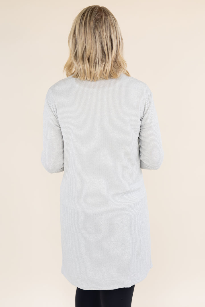 A woman viewed from behind wearing a light gray ribbed knit long-sleeve tunic top, set against a solid beige studio background.
