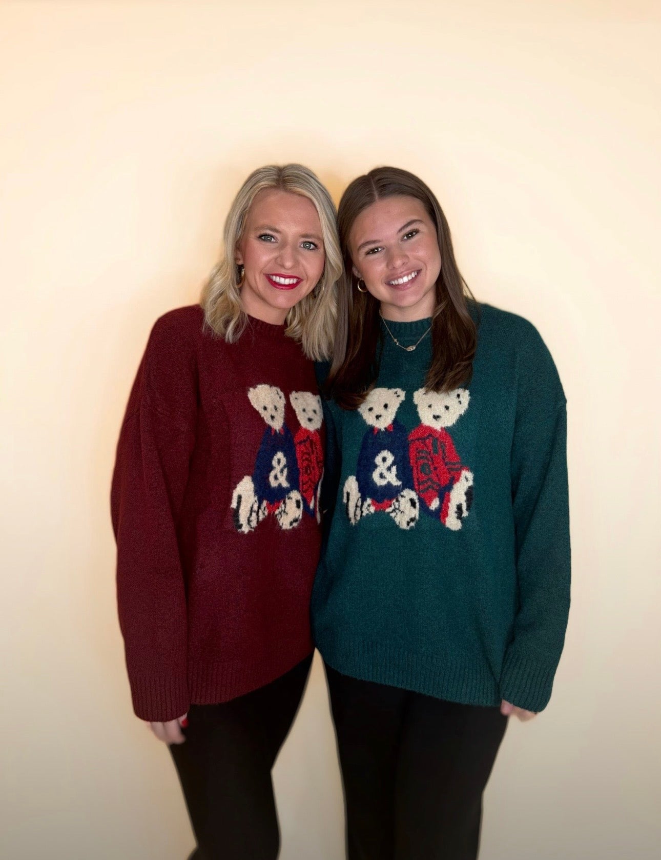 Two women wearing holiday teddy bear sweaters in burgundy and forest green, paired with black pants, standing together against a neutral background.