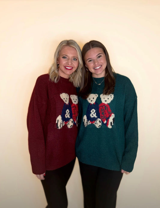 Two women wearing holiday teddy bear sweaters in burgundy and forest green, paired with black pants, standing together against a neutral background.