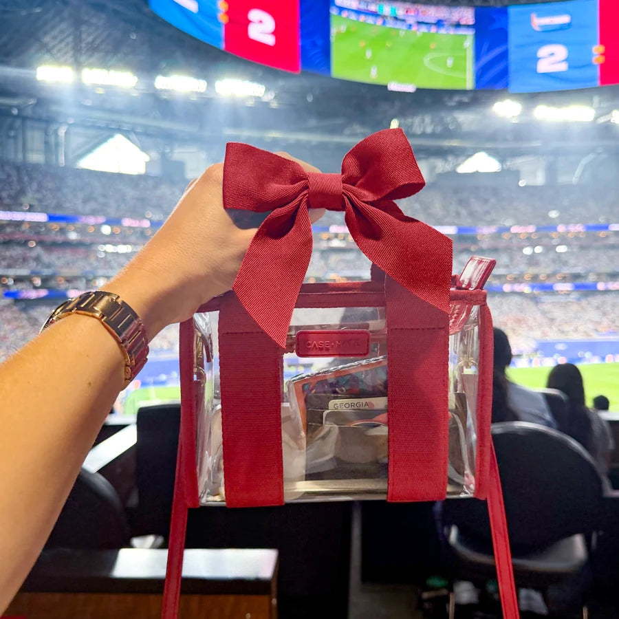 Case-Mate Varsity Red Bow Jelly Stadium Bag held inside a football stadium, featuring a clear stadium-approved design, red bow and essentials inside.
