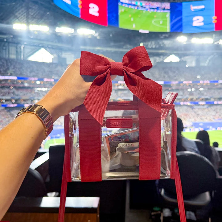 Case-Mate Varsity Red Bow Jelly Stadium Bag held inside a football stadium, featuring a clear stadium-approved design, red bow and essentials inside.