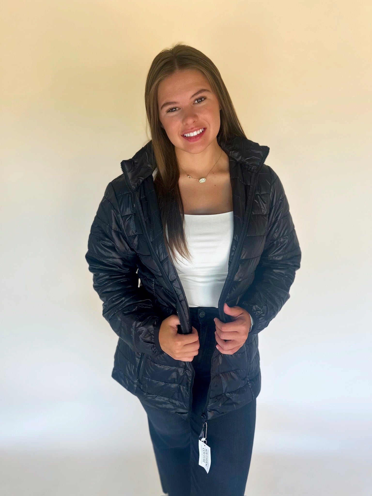 Woman wearing a black jacket over a white top and dark pants on a plain background