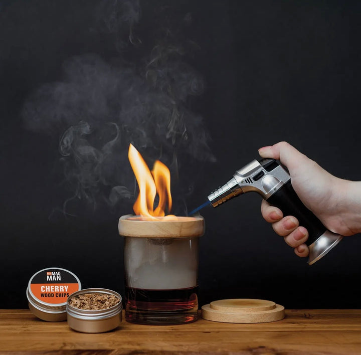 Hand using the Mad Man Whiskey Smoker torch to infuse a whiskey cocktail with rich smoke using cherry wood chips and a wooden smoker lid.