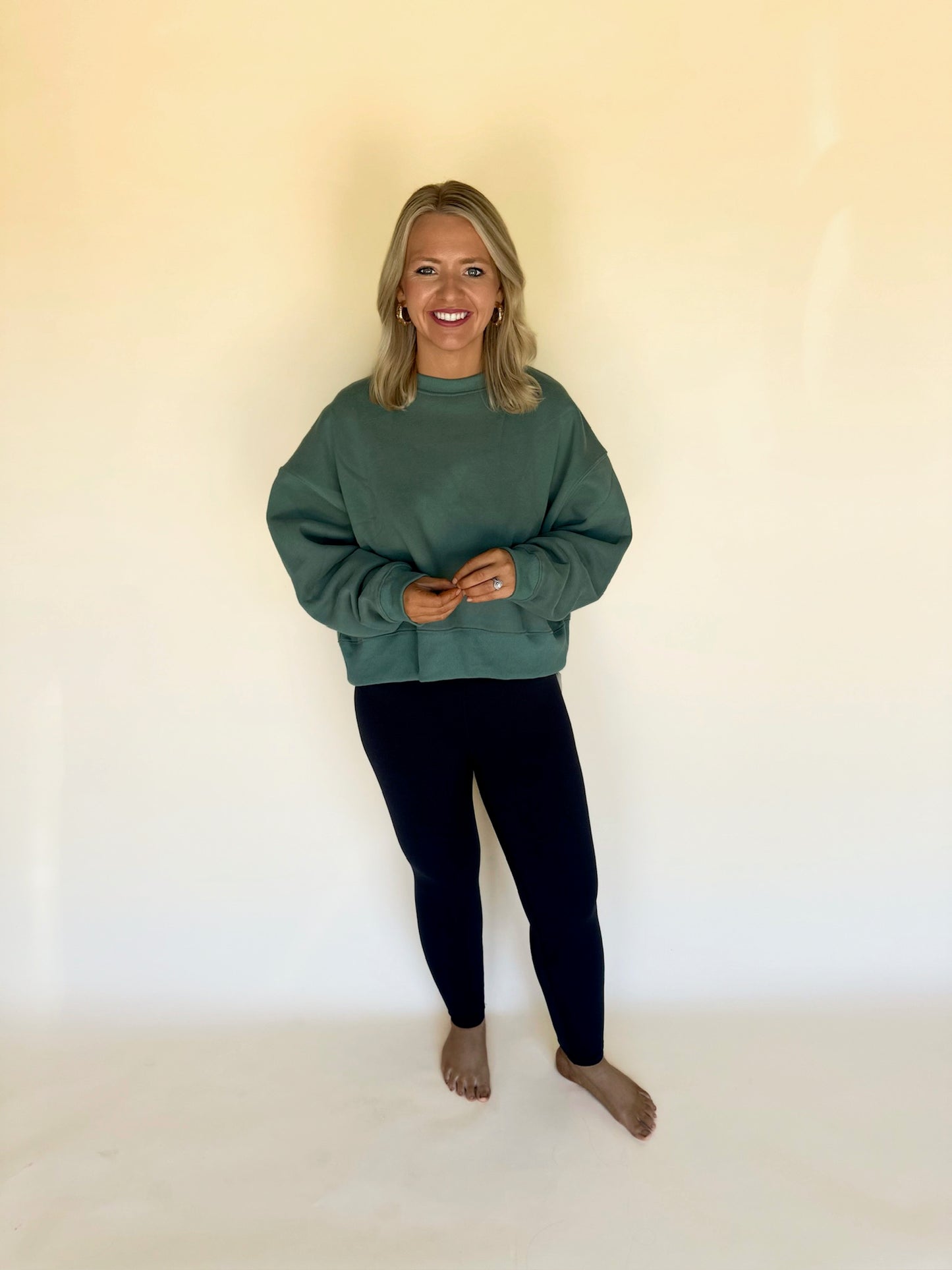 Person wearing a green sweatshirt and black leggings standing against a white wall.