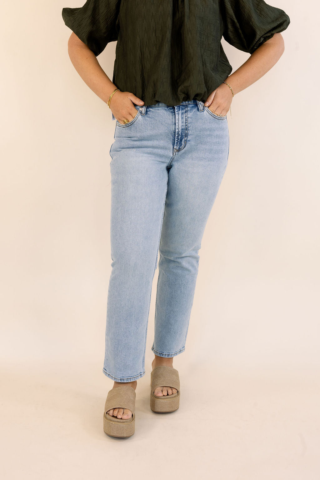 A model wearing light-wash blue jeans, a dark green short-sleeve blouse, and taupe platform sandals, standing against a solid light beige background.