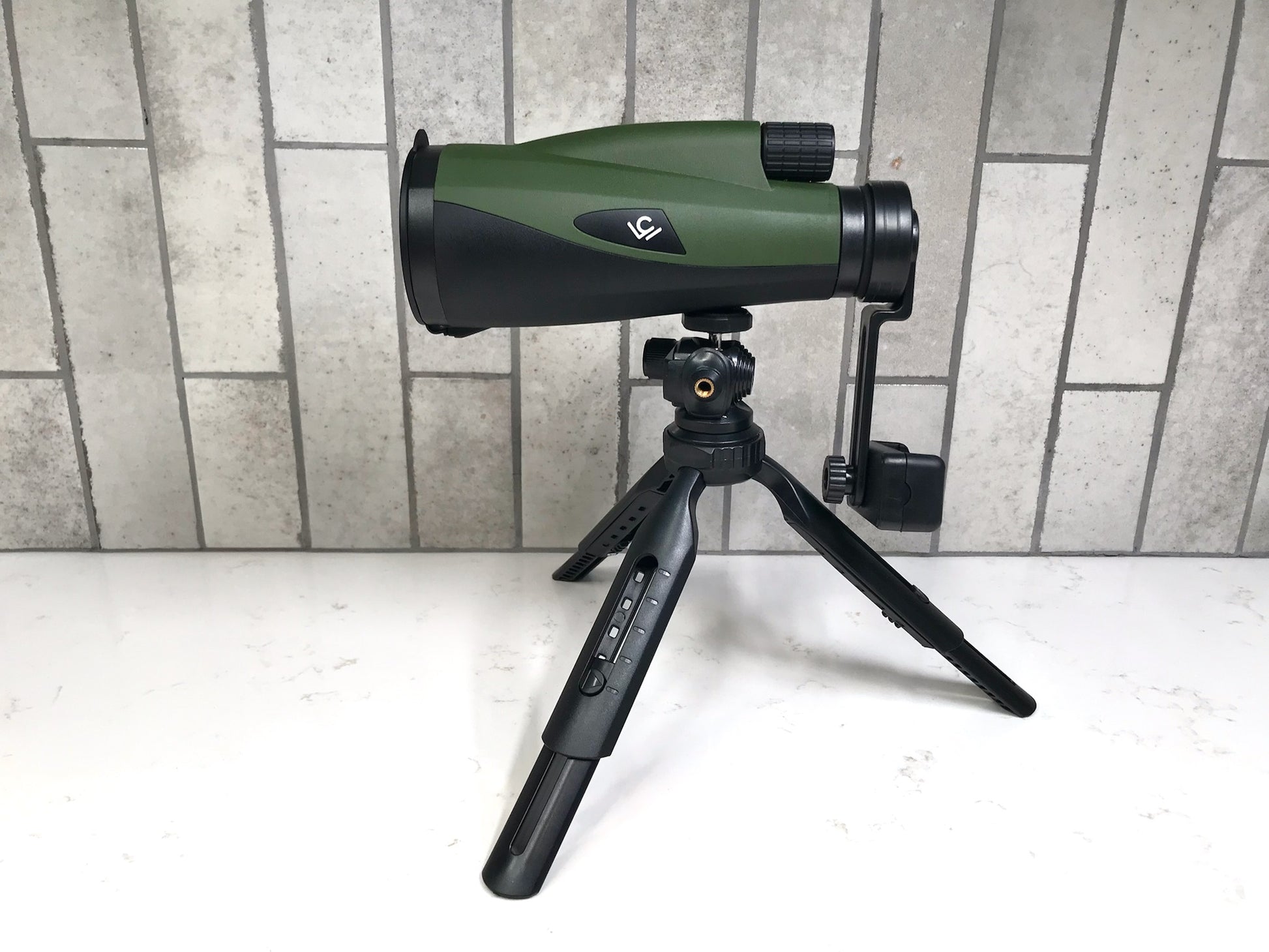 Green and black spotting scope on a tripod against a tiled wall.